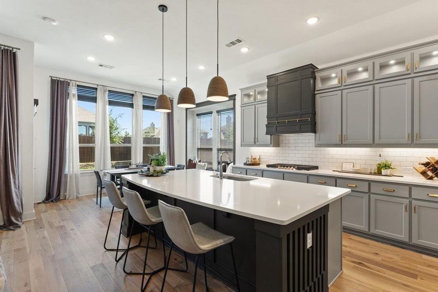 Kitchen featuring light wood-type flooring, a kitchen island with sink, stainless steel gas stovetop, backsplash, and pendant lighting