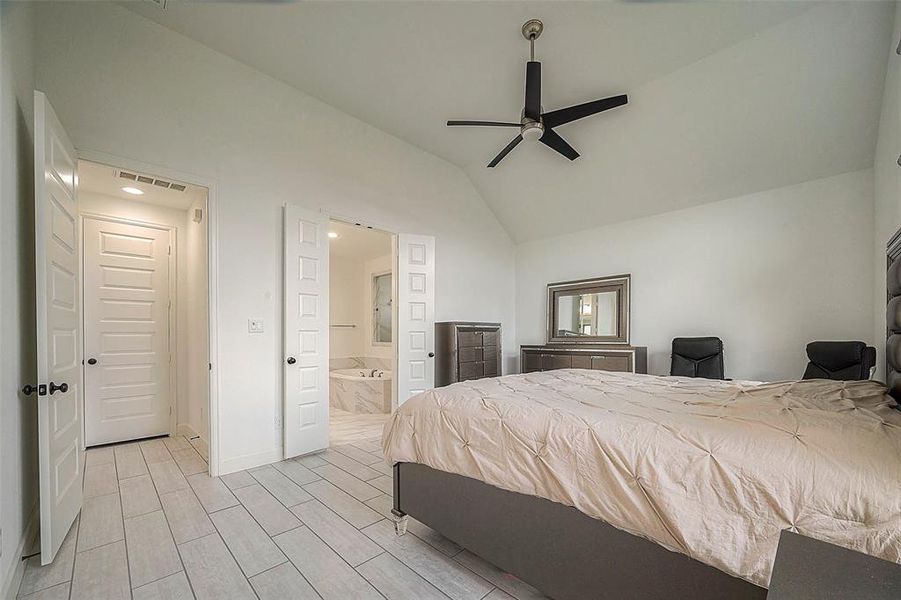 This is a spacious bedroom featuring a high vaulted ceiling with a modern fan, light gray tile flooring, and ample natural light. The room includes a large bed, matching furniture, and direct access to an en suite bathroom.