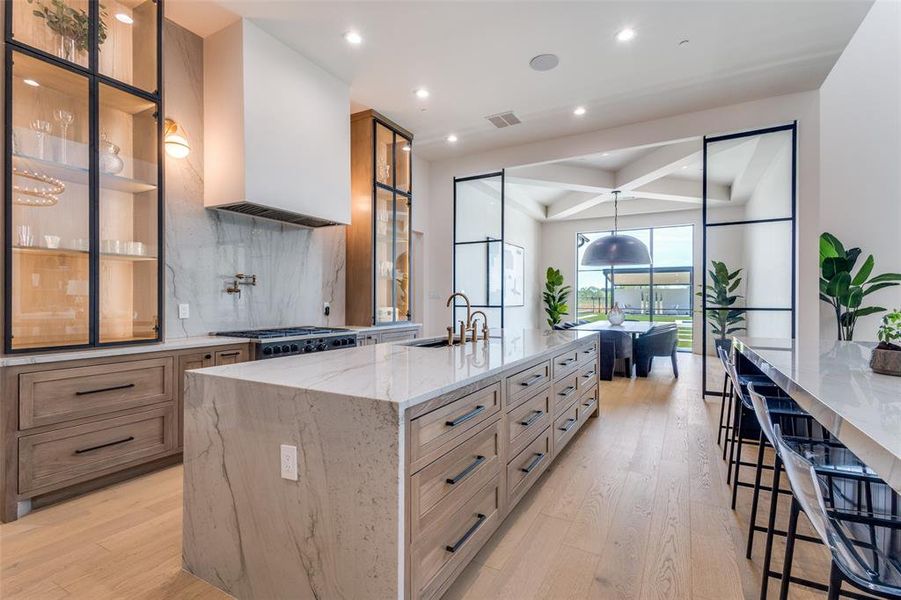 Kitchen with hanging light fixtures, light wood-type flooring, a center island with sink, light stone counters, and tasteful backsplash