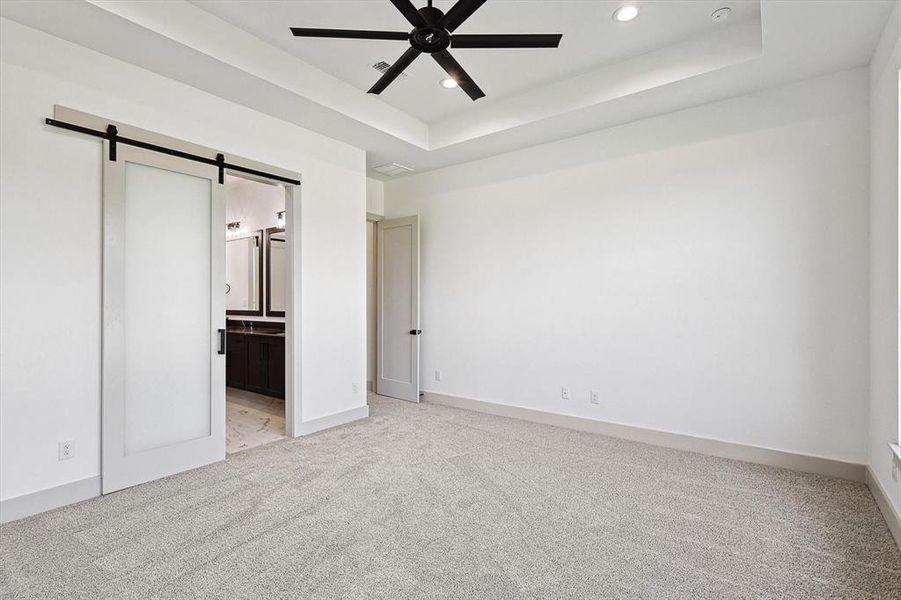 Unfurnished bedroom with light carpet, ensuite bath, ceiling fan, a tray ceiling, and a barn door