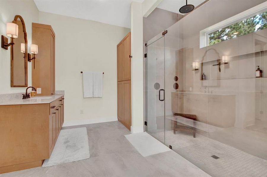 The modern primary bathroom features a spacious walk-in shower with a frameless glass enclosure, complemented by sleek wooden vanities with marble countertops, matching wooden storage cabinets, and stylish wall-mounted light fixtures.