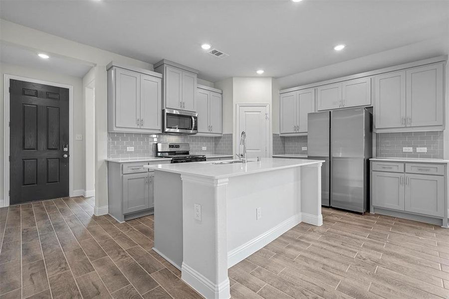 Kitchen with appliances with stainless steel finishes, gray cabinetry, a center island with sink, decorative backsplash, and light wood-type flooring