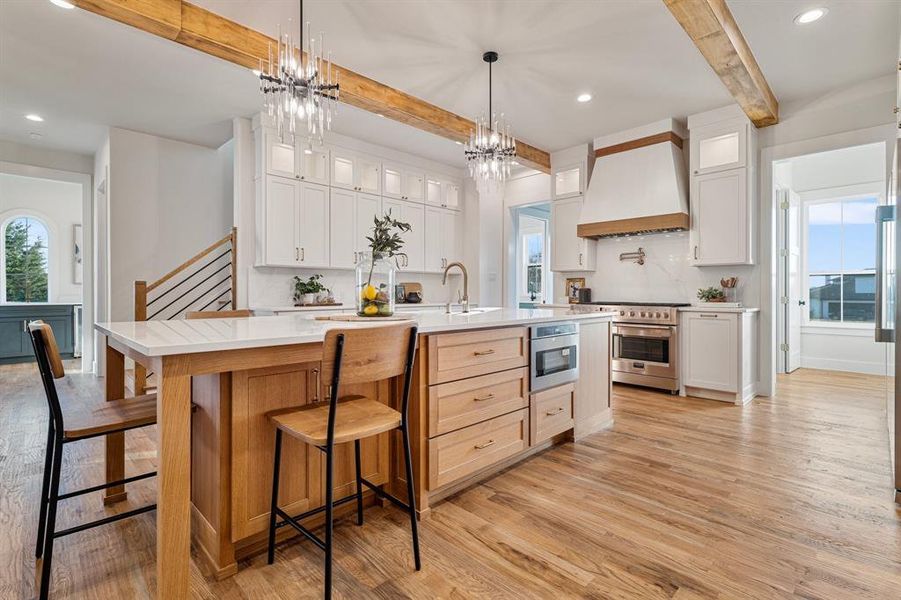 Kitchen featuring custom exhaust hood, beam ceiling, light wood-type flooring, range with two ovens, and a kitchen island with sink