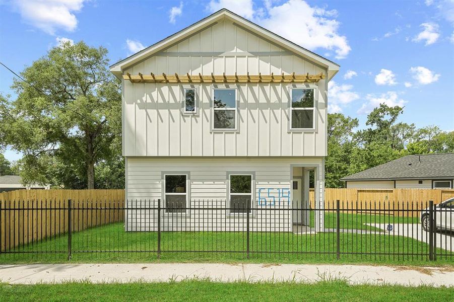 This is a two-story home with a modern farmhouse aesthetic, featuring a light exterior, contrasting trim, and a covered porch. The property is enclosed by a black metal fence and includes a grassy front yard with a mature tree in the vicinity.