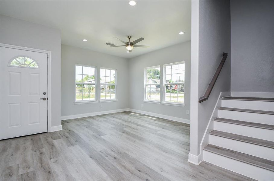 This is a bright, spacious entryway main living room and with modern gray tones, wood-look flooring, and plenty of natural light from large windows. There is a staircase to the right, leading to the upper level.