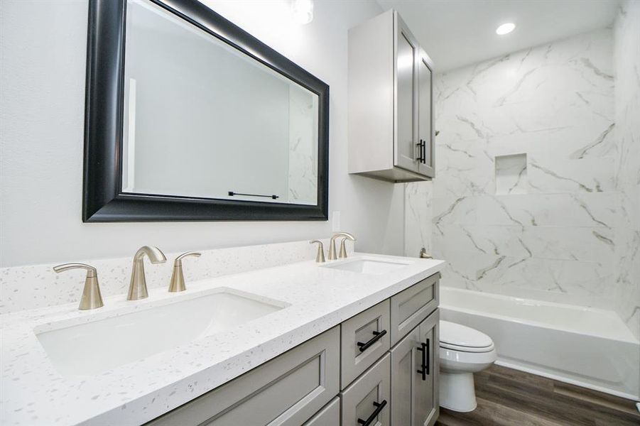 This is a modern secondary bathroom featuring a double vanity with brushed nickel fixtures, white countertops, and a large mirror. The space includes a tub-shower combo with marble-like wall tiles, a toilet, and grey cabinetry offering ample storage. The flooring has a wood-look finish.