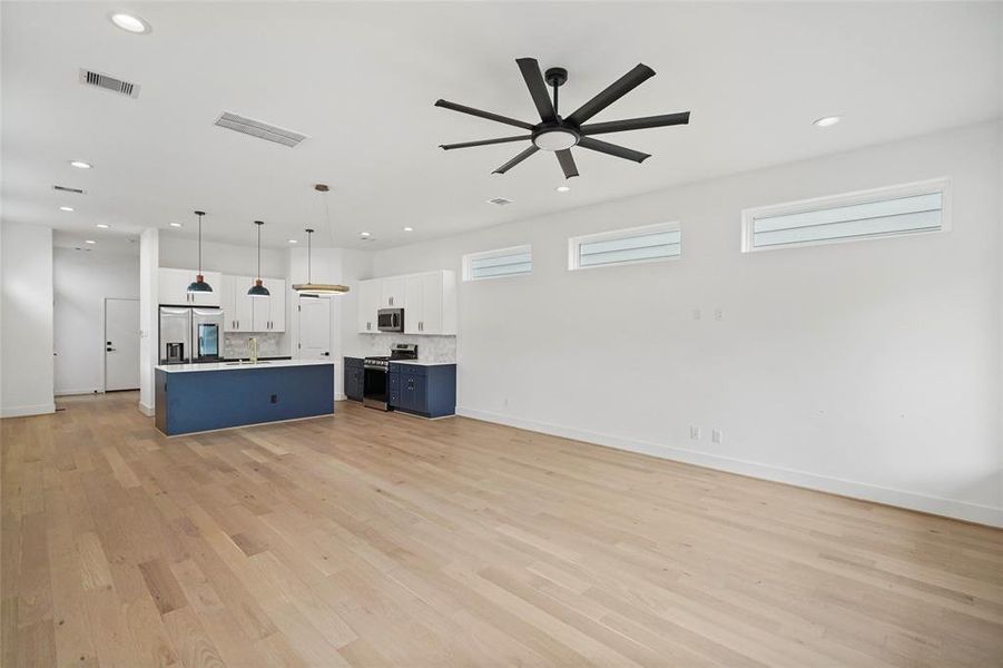 This is a modern open-concept living space with a bright kitchen featuring white cabinetry, stainless steel appliances, and a contrasting blue center island. The room is well-lit with natural light from horizontal windows and includes a contemporary ceiling fan. The flooring throughout is white oak hardwood.