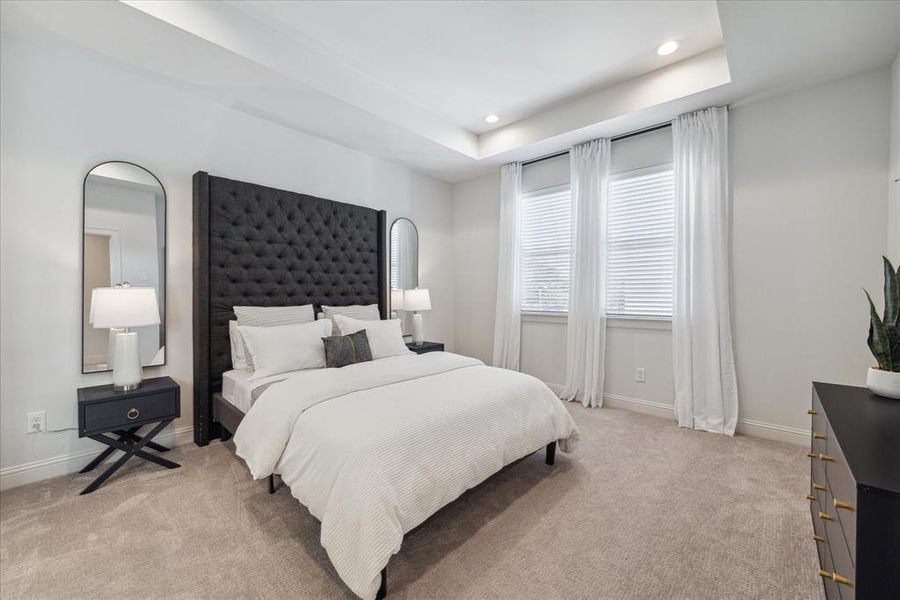 This spacious primary bedroom offers a serene retreat with soft carpeting and large windows that flood the room with natural light. The recessed ceiling and neutral color palette create a soothing and airy ambiance, perfect for relaxation.