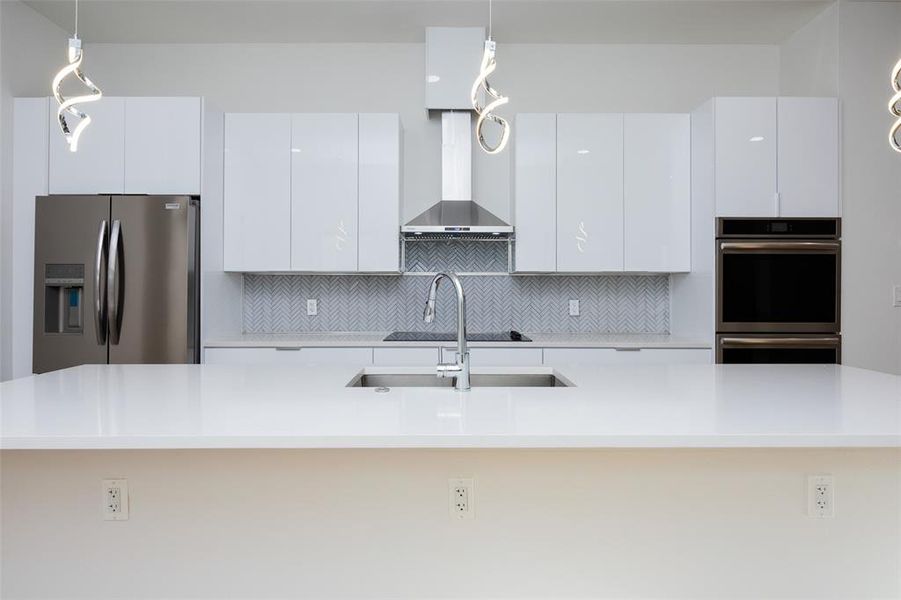 Kitchen with wall chimney range hood, stainless steel appliances, a kitchen island with sink, backsplash, and pendant lighting