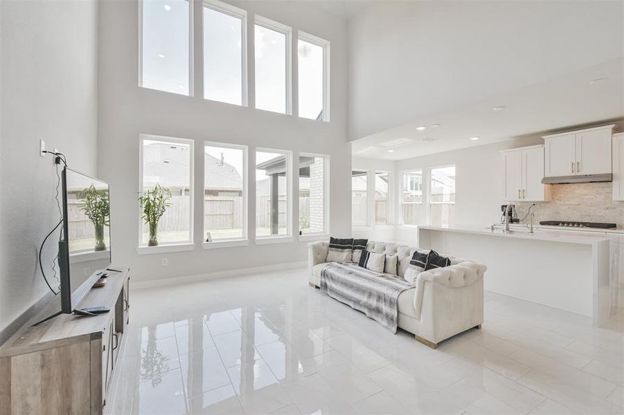 You're looking at a bright, modern open-plan living space featuring high ceilings, large windows, and a sleek kitchen with updated appliances. The area is bathed in natural light, enhancing the spacious feel and highlighting the glossy tile flooring.