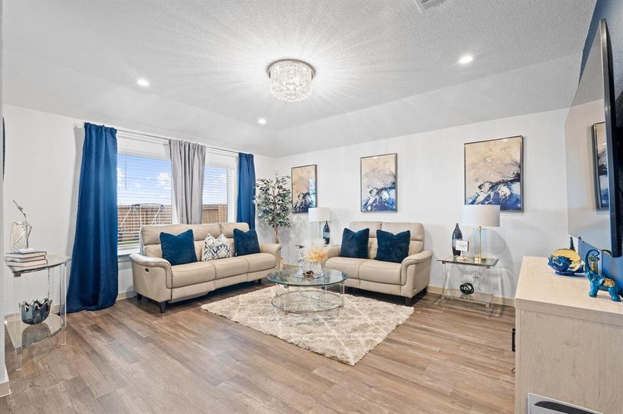 This is a modern and well-lit living room featuring a neutral color palette with bold blue accents. It includes comfortable seating, and a large window that allows for plenty of natural light. The room is finished with premium laminate  flooring and a stylish chandelier. Approximate Measurements: 16x14