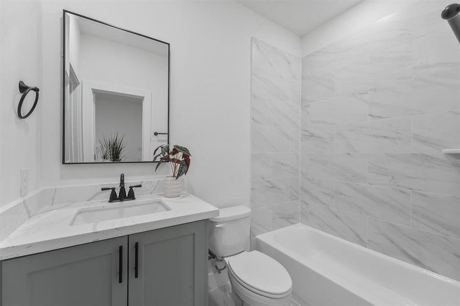 Modern secondary bathroom featuring a faux marble tile, a modern sleek gray custom vanity with a quartz countertop, custom modern framed matte black mirror, and a full-size bathtub surrounded by stone tiles.