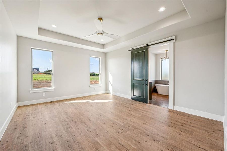 Unfurnished bedroom featuring ceiling fan, hardwood / wood-style floors, a barn door, and a tray ceiling