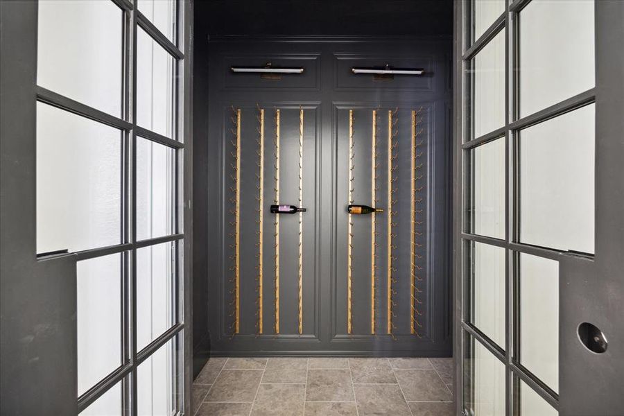 The iron double doors open to the chic wine cellar. The wine cellar features tile flooring, sleek gold wine racks and wall-mounted sconces.