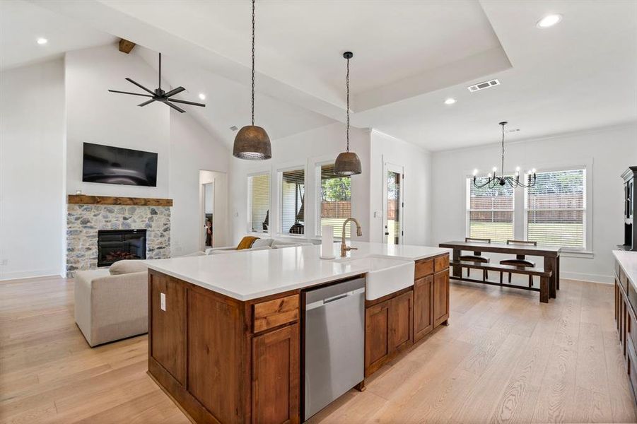 Kitchen with chandelier, dishwasher, a stone fireplace, light hardwood floors, and an island with sink