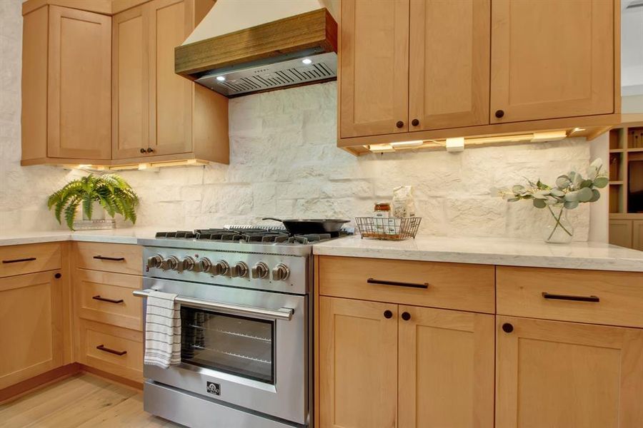 This is a modern kitchen with light wood cabinetry, stainless steel appliances, and a textured white stone backsplash. The countertops are a light quartz, and under-cabinet lighting adds warmth to the space.