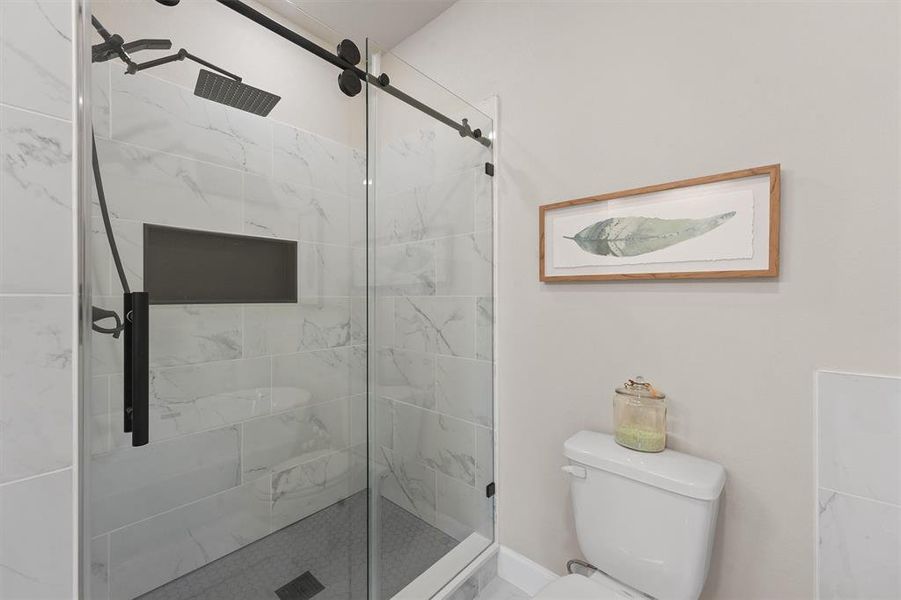 This is a modern bathroom featuring a walk-in shower with marble-like tiles and black fixtures, a toilet, and minimalistic decor with a framed artwork on the wall.