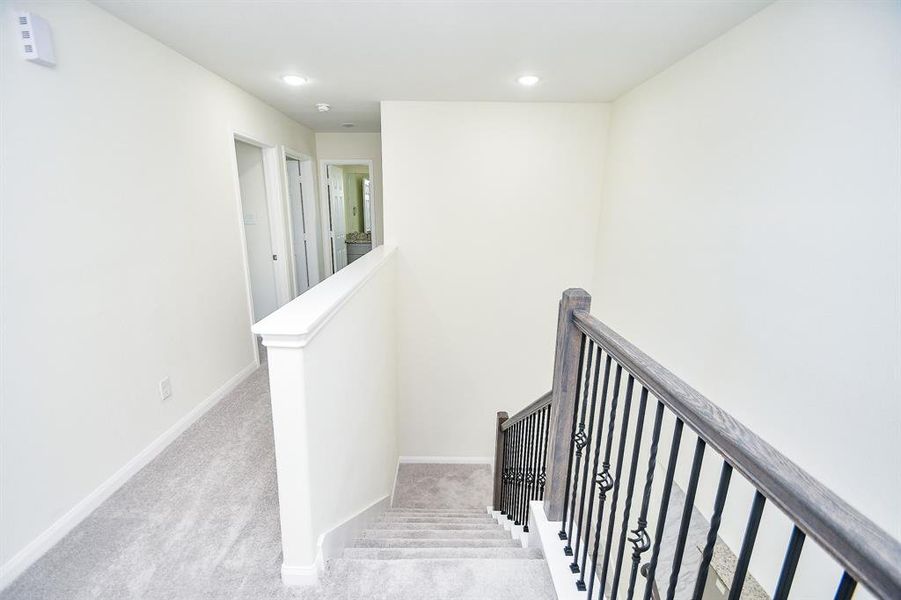 Interior of a house showing a carpeted hallway leading to stairs with a white balustrade and dark wooden banisters.