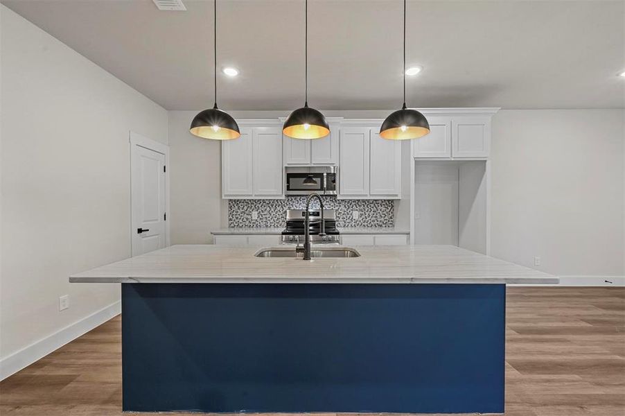 Kitchen with hanging light fixtures, an island with sink, white cabinets, light stone countertops, and sink