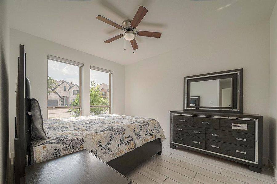 This is a bright, modern bedroom featuring large windows for plenty of natural light, a ceiling fan for comfort, and stylish hardwood flooring. The room is well-appointed with a large bed and a contemporary dresser with a mirror, providing a sleek and comfortable living space.