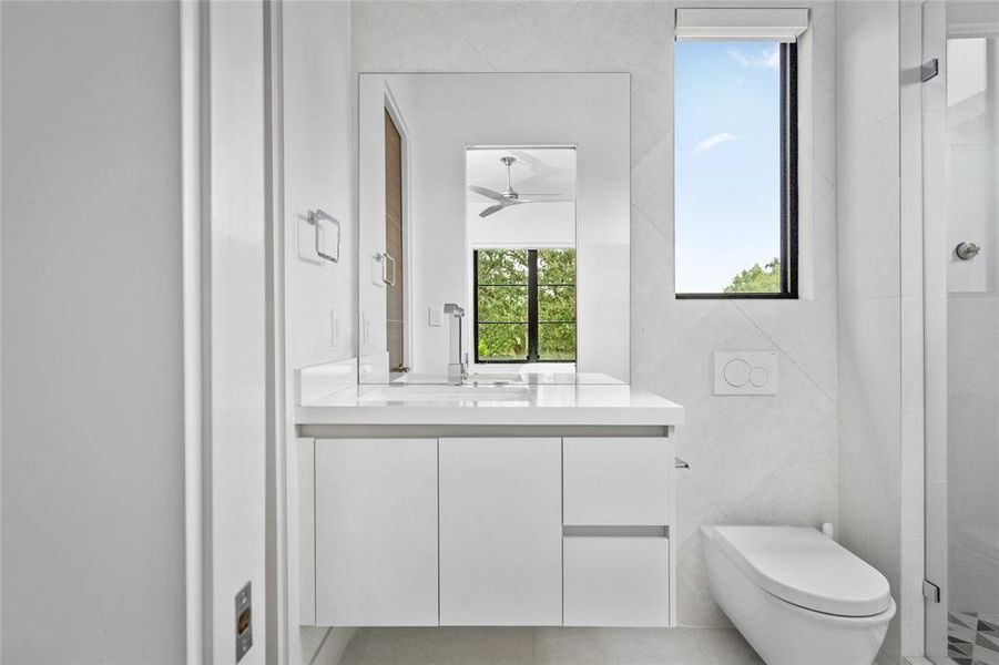 This secondary ensuite bathroom features sleek, modern fixtures with a clean white vanity and chrome faucet. The walk-in shower is enhanced by geometric-patterned floor tiles and a frameless glass enclosure. Natural light flows through the high windows, creating a bright and airy atmosphere.