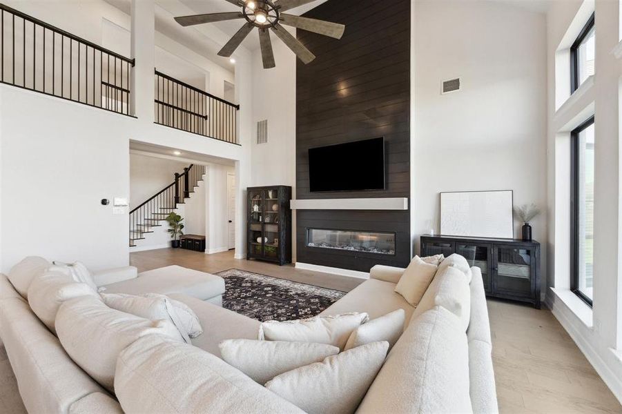 Another view of the living area with a dark wood accent wall, fireplace, abundant natural light from tall windows, and an open staircase leading to an upper level. Perfect for entertaining.