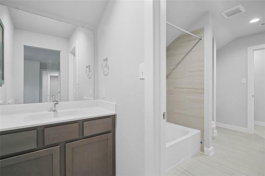 This secondary jack-n-jill bath features tile flooring, bath/shower combo with tile surround, stained wood cabinets, beautiful light countertops, mirror, sleek fixtures and modern finishes.