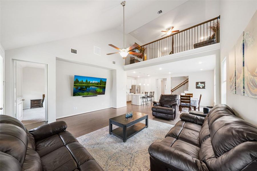 This image shows a spacious, well-lit living area with high ceilings, featuring a modern open-concept design that connects to a dining space and kitchen. The room is equipped with a ceiling fan and has a second-floor balcony overlook, providing a sense of openness and connectivity within the home.