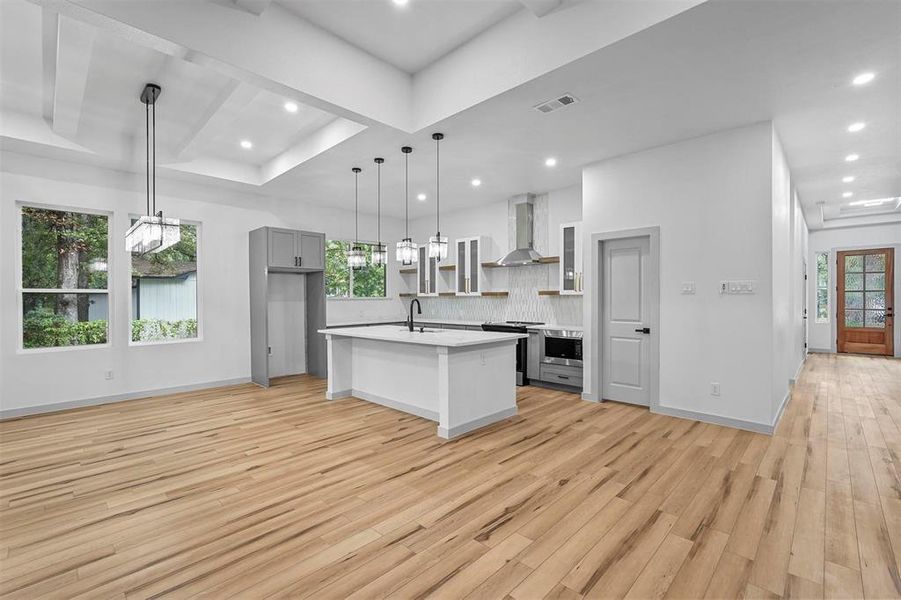 Kitchen with light wood-type flooring, decorative light fixtures, gray cabinets, wall chimney range hood, and an island with sink