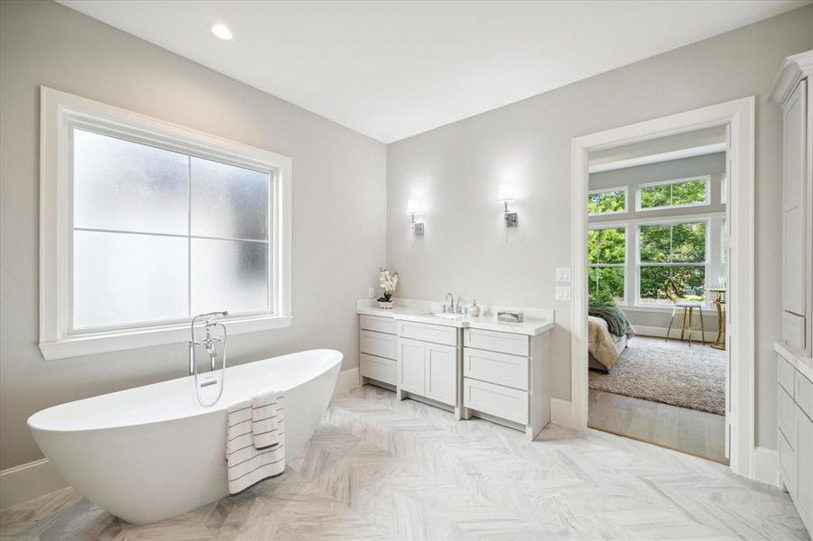 This is a spacious and modern bathroom featuring a standalone bathtub, double vanity with ample storage, frosted window for privacy, elegant lighting fixtures, and herringbone patterned flooring. There's also a view into an adjoining room with natural light and a view of greenery.