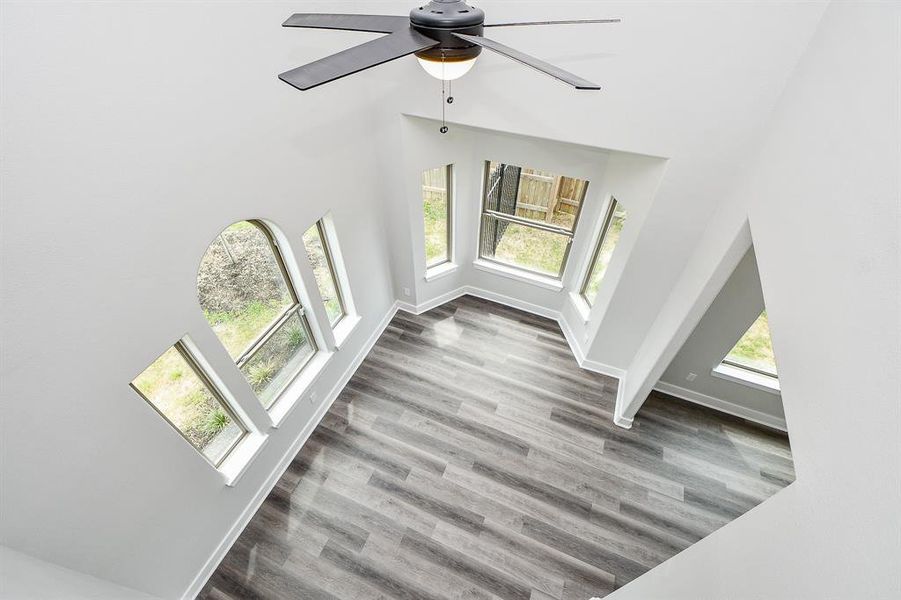 This is a bright, modern room with high ceilings, a unique geometric design, large windows, and stylish wood-look flooring. It features a contemporary ceiling fan and offers a spacious, airy feel.