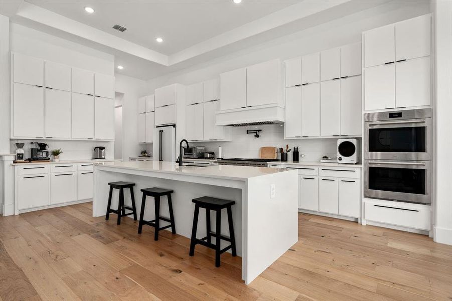 Delightful kitchen featuring light hardwood / wood-style floors, stainless steel appliances, an island with sink, white crisp cabinets, and a raised ceiling