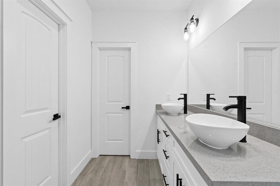 This is a modern bathroom featuring a sleek double vanity with above-counter basins, black faucets, and a light granite countertop. The space is accented with black hardware, contrasting with the clean white walls and cabinetry, and complemented by light wood-look flooring.