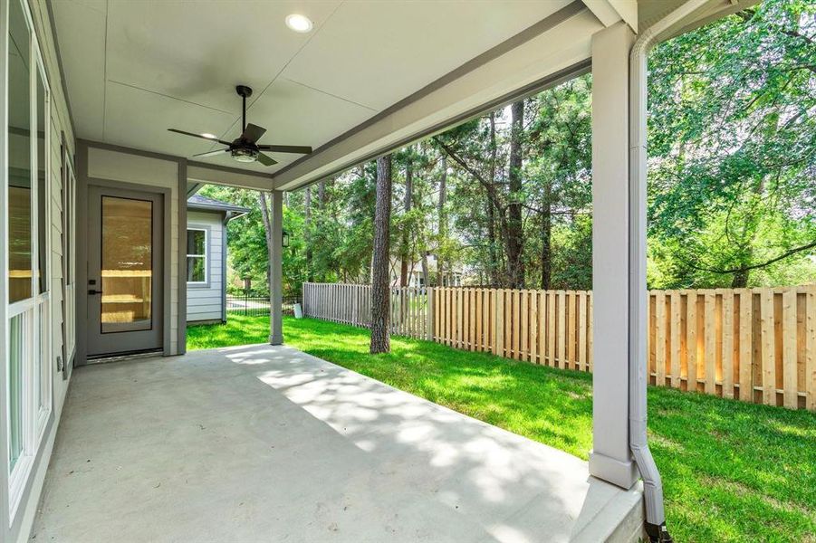 A covered patio with a ceiling fan, opening to a fenced in backyard with beautiful surrounding trees that brings in lots of shade.