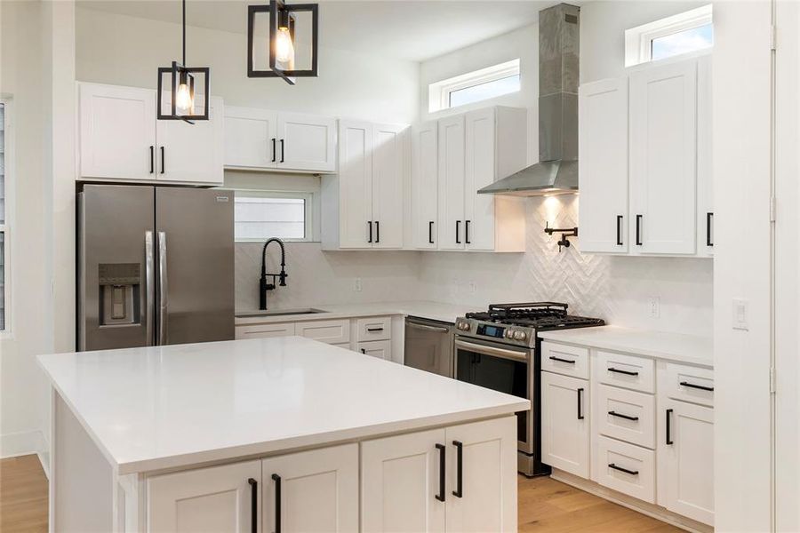 This modern kitchen features white shaker-style cabinetry, stainless steel appliances, a gas stove, and a central island with a quartz countertop. The space is lit by stylish pendant lights and natural light from the high windows above the backsplash.