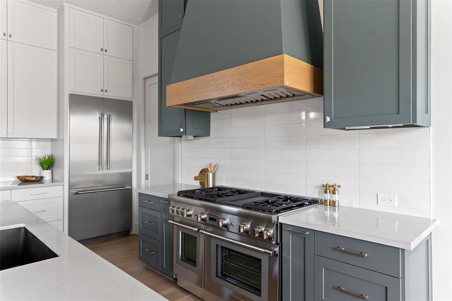 This is a modern kitchen featuring stainless steel appliances, white subway tile backsplash, and a mix of white and teal cabinetry with sleek hardware. It includes a gas range with a wood-trimmed hood vent and light quartz countertops.