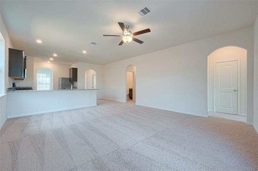 Spacious empty room with carpet, ceiling fan, and partial view of a kitchen with appliances. Bright, clean interior with windows and doors.