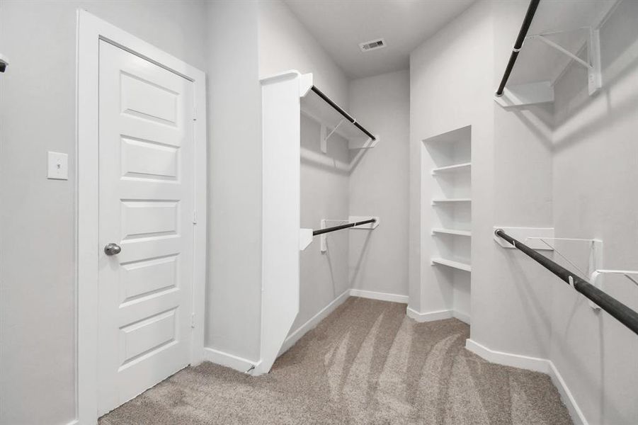 Expansive walk-in closet, adorned with high ceilings and luxurious carpet. Immerse yourself in the inviting atmosphere created by warm paint tones, utilize the convenience of built-in shelving, and experience the modern and functional retreat accentuated by dark finishes. Sample photo of completed home. Actual colors and selections may vary.