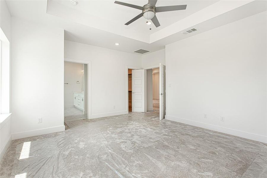 Unfurnished bedroom with light colored carpet, ensuite bath, ceiling fan, and a tray ceiling