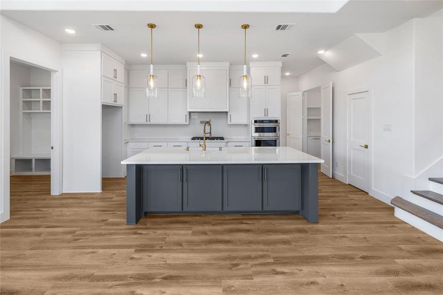 Kitchen with light wood-type flooring, an island with sink, stainless steel double oven, white cabinetry, and hanging light fixtures