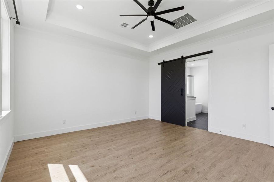 Unfurnished bedroom with light wood-type flooring, a barn door, ensuite bath, a raised ceiling, and crown molding