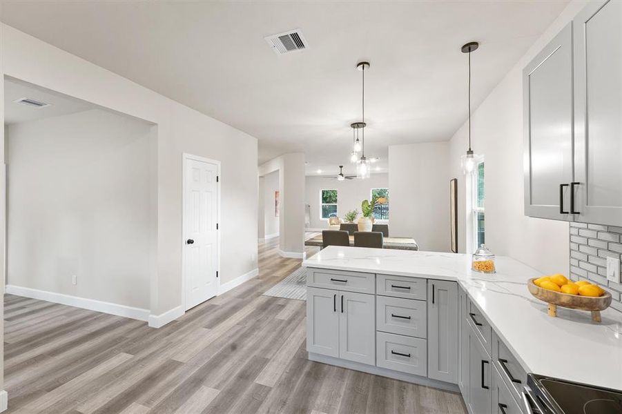 Modern kitchen with gray cabinetry, stainless steel appliances, white quartz countertops, and subway tile backsplash. Open plan view into a bright living space with contemporary wood-look flooring.