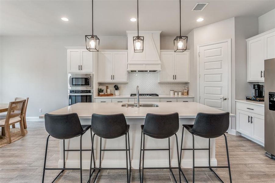 Kitchen featuring custom range hood, tasteful backsplash, a kitchen island with sink, stainless steel appliances, and light hardwood / wood-style floors
