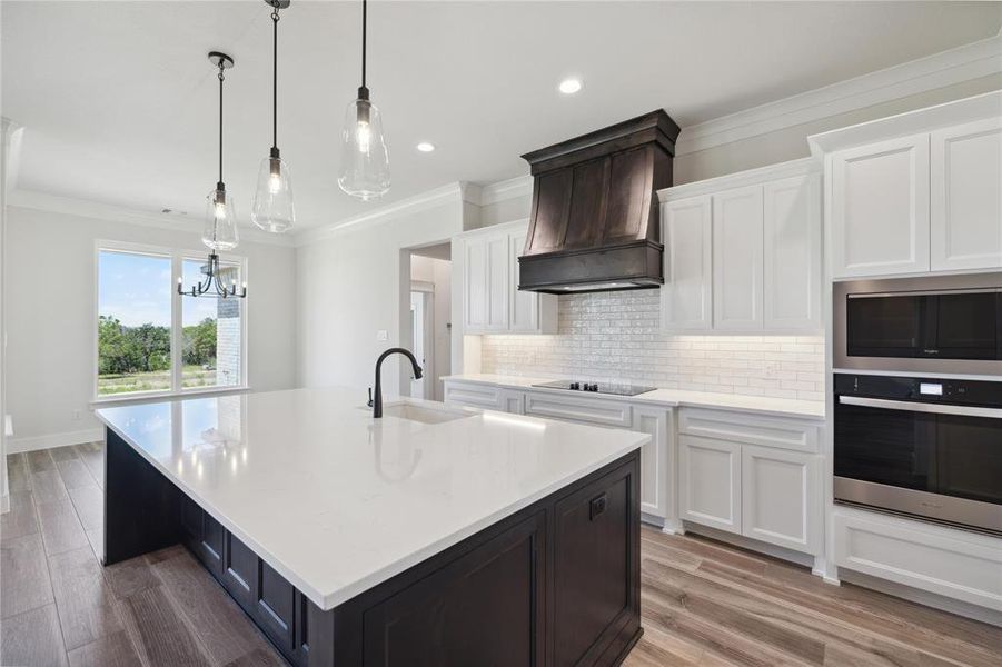 Kitchen featuring white cabinets, decorative backsplash, custom range hood, light hardwood / wood-style floors, and a kitchen island with sink