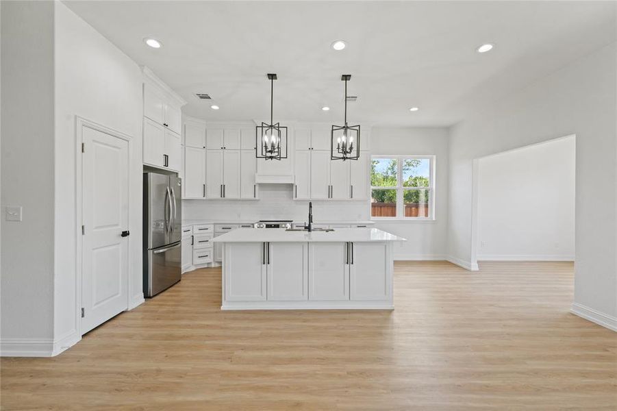 Kitchen with light hardwood / wood-style floors, pendant lighting, white cabinets, a kitchen island with sink, and stainless steel refrigerator
