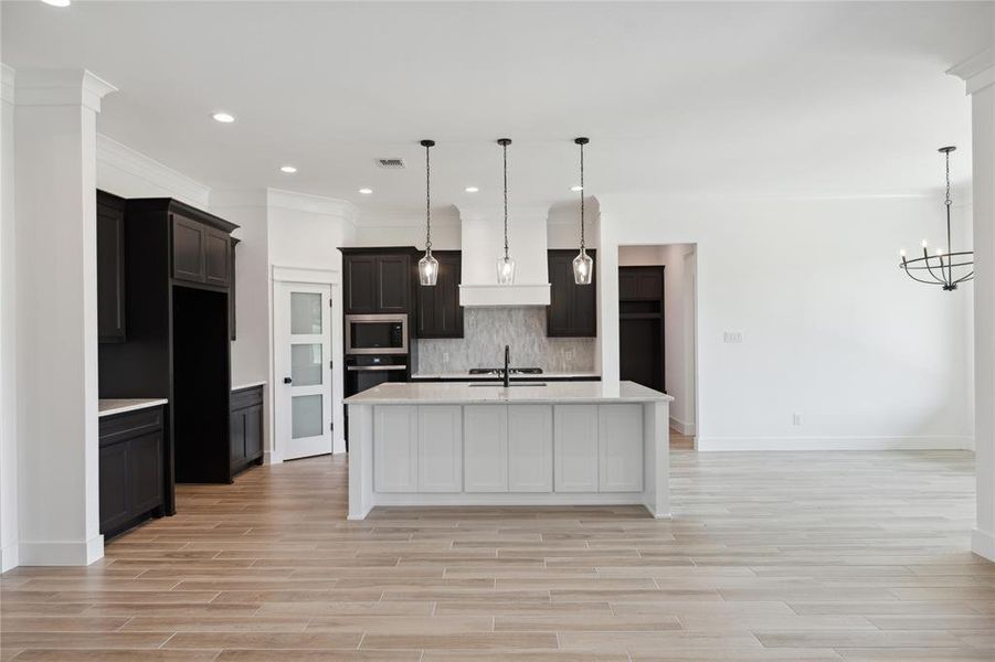 Kitchen with light wood-type flooring, decorative light fixtures, appliances with stainless steel finishes, decorative backsplash, and a kitchen island with sink