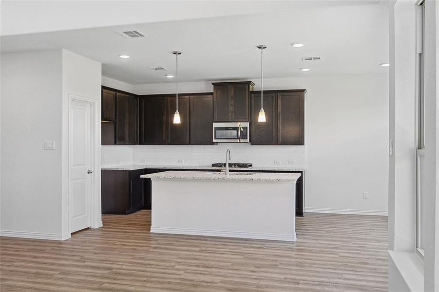 Kitchen with hanging light fixtures, backsplash, dark brown cabinetry, light wood-type flooring, and an island with sink