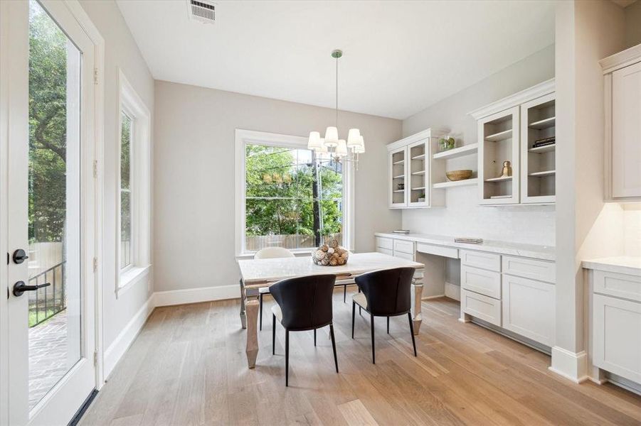 This is a bright, contemporary space featuring a dining area with a modern table and chairs, built-in white cabinetry, and ample natural light from large windows and a glass door leading to the outside. The room has light hardwood floors and a fresh, neutral color palette.