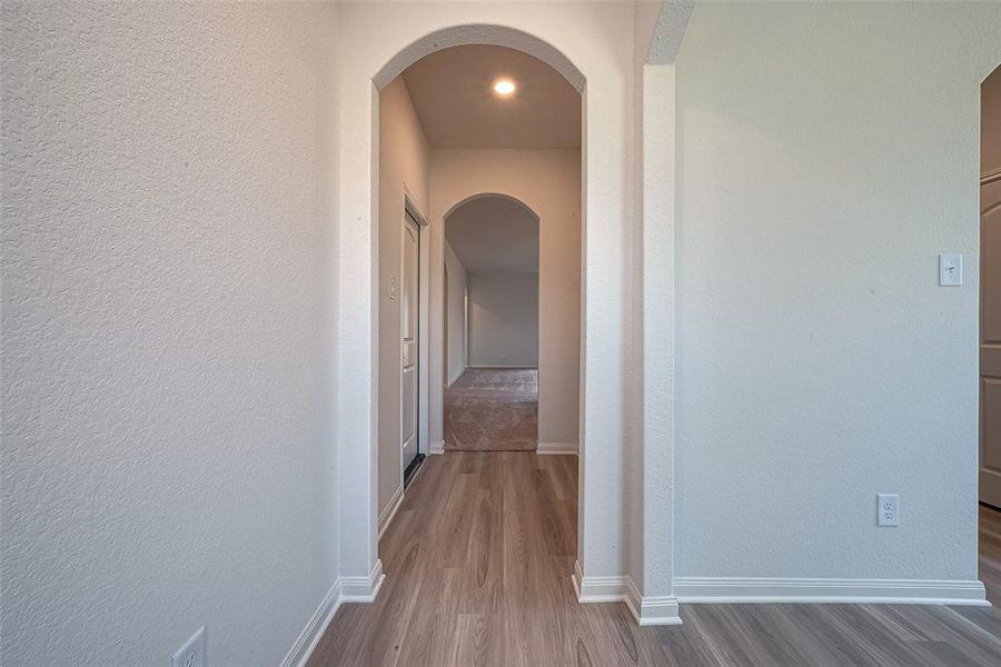 An hallway with wooden floors, white walls, and an arched entry leading to another room.