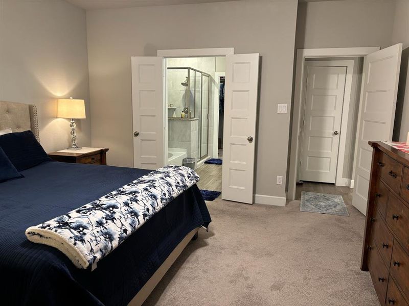 This is a well-appointed bedroom featuring two white doors leading to a bathroom with a visible shower and a closet, neutral carpet. The room has a calm, neutral color palette.